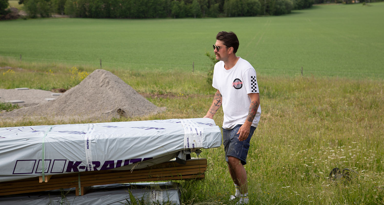 Man i vit t-shirt och shorts lyfter presenningklädd trälast på grön sommaräng.