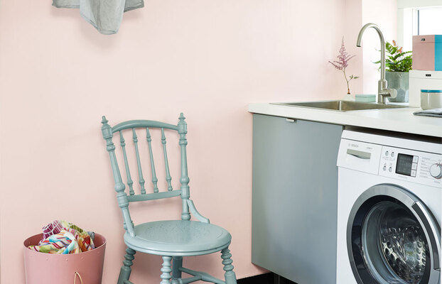 Tvättstuga med pastellrosa vägg, mintgrön stol, frontmatad tvättmaskin, diskho och klädkorg.