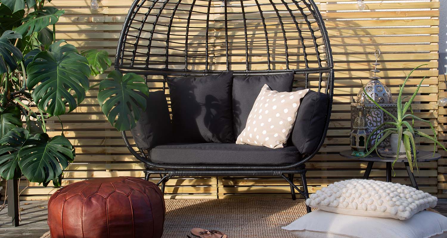 Uteplats med svart rottingsoffa, beige prickkudde, röd läderpuff, monstera, ljusslinga.