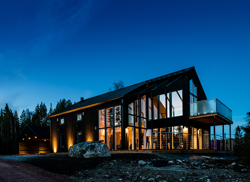 Modern tvåvåningsvilla med glasfasad och varma ljus mot blå kvällshimmel i skogsmiljö.