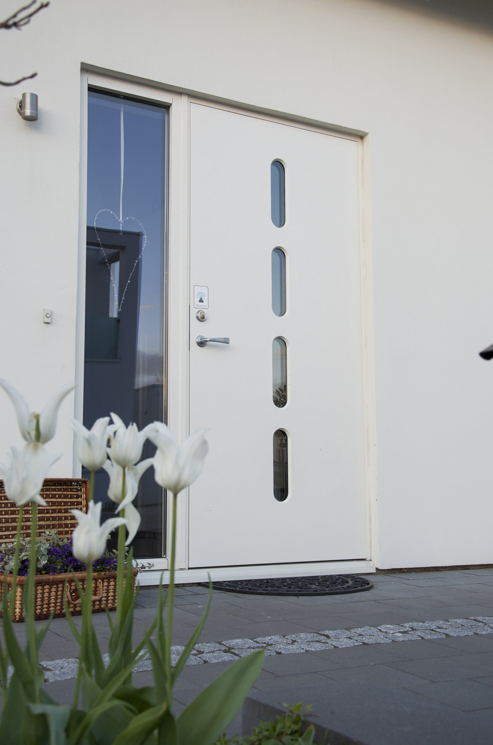 Vit entrédörr med fyra smala fönstergluggar och hängande hjärta, vita tulpaner.