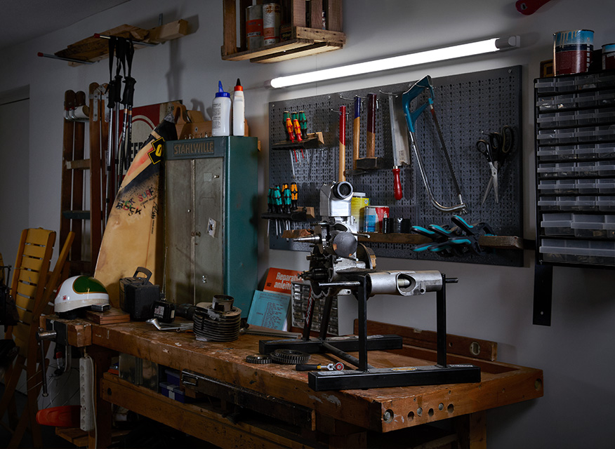Mörk träbänk med metallsvarv, upplyst pegbord med skruvmejslar, blågrönt skåp och hjälm