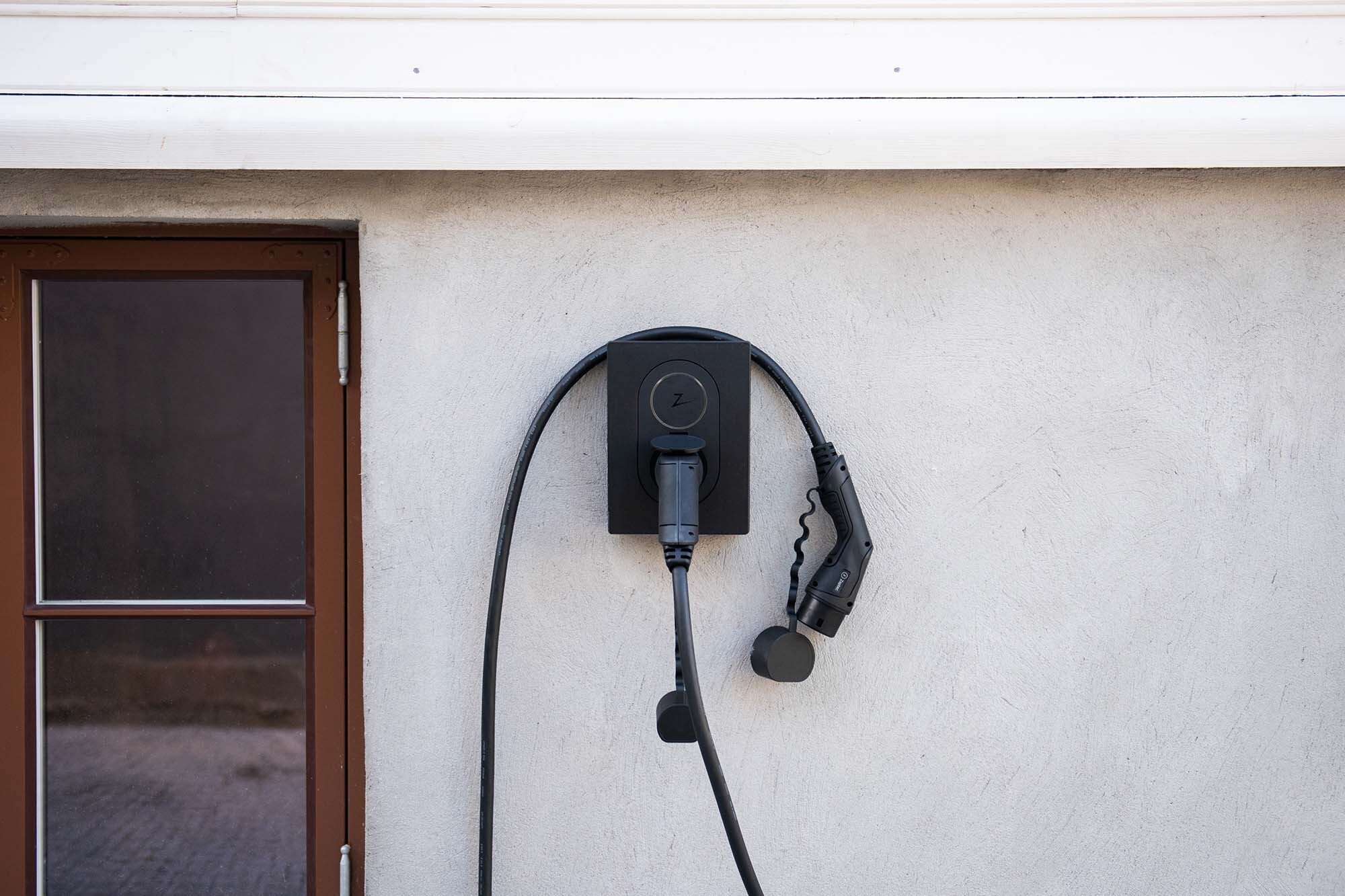 Svart elbilsladdare med kabel och kontakt på ljusgrå vägg bredvid brunt fönster