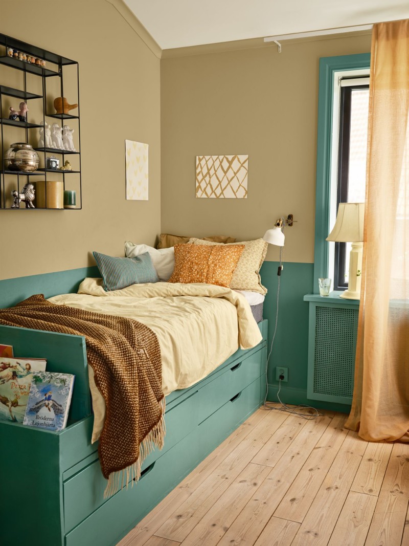 Mysigt hörnrum med turkos dagbädd, beige väggar, gula lakan, orange kuddar och fönster.