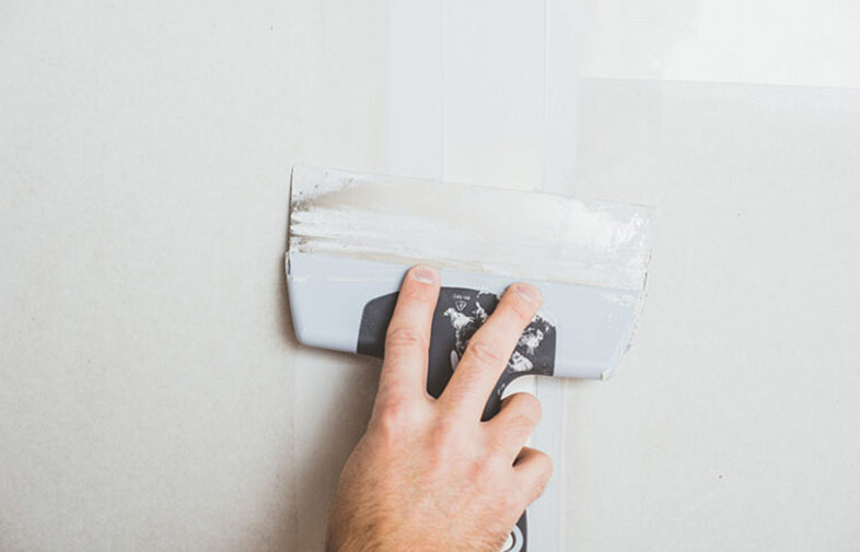Hand med spackelspade jämnar ut vit spackelmassa på ljusgrå vägg.