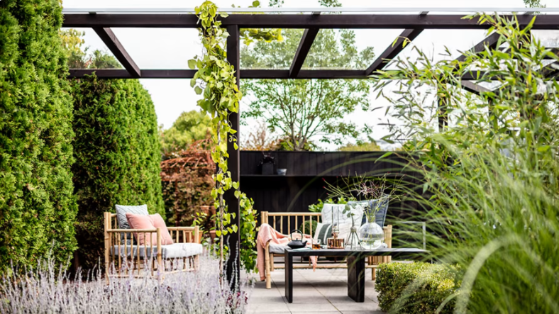 Modern uteplats med svart pergola, ljusgrå rottingsoffor med rosa kuddar, grön växtmiljö