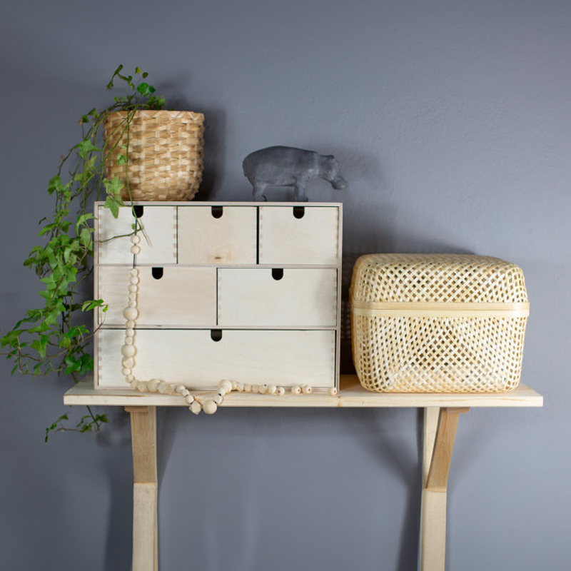 Ljus träbyrå med sex lådor, hängande grön växt, grå flodhästfigur och rottingkorg mot grå vägg.