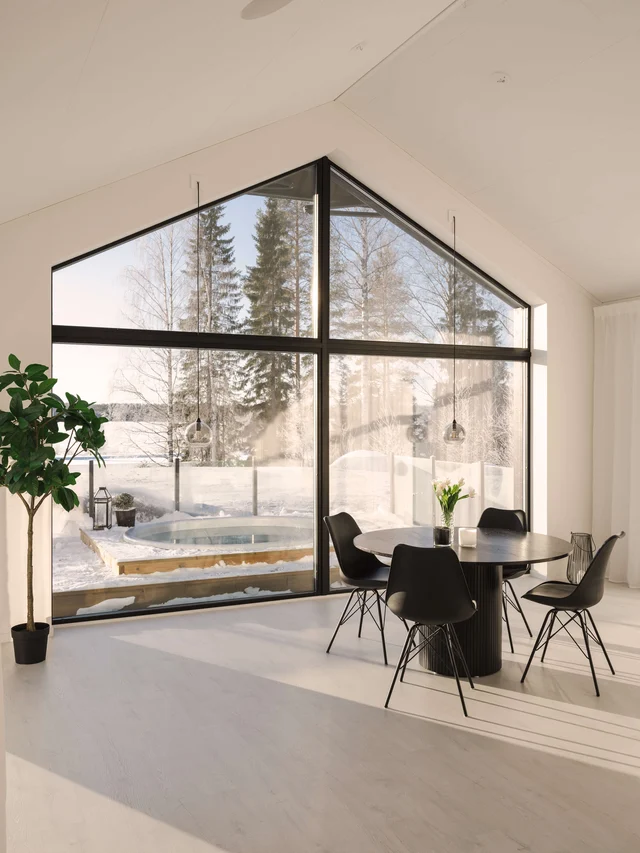 Ljust vardagsrum med svart runt bord och stolar vid stora panoramafönster mot snötäckt skog
