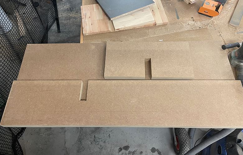 Flera ljusbruna MDF-skivor med urtag liggande på arbetsbänk i träverkstad, verktyg runtom.