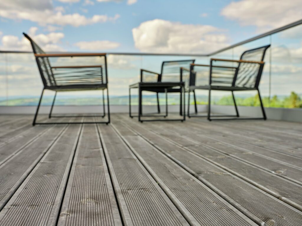 Uteplats med grå trall, glasräcke mot vy, svart bord och fyra stolar under molnig himmel.