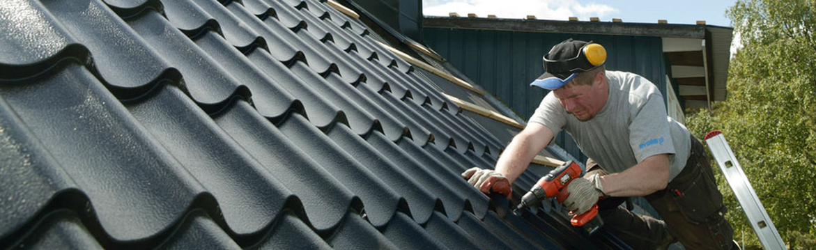 Man på mörkgrått plåttak fäster plåt med orange borrmaskin vid blå husvägg