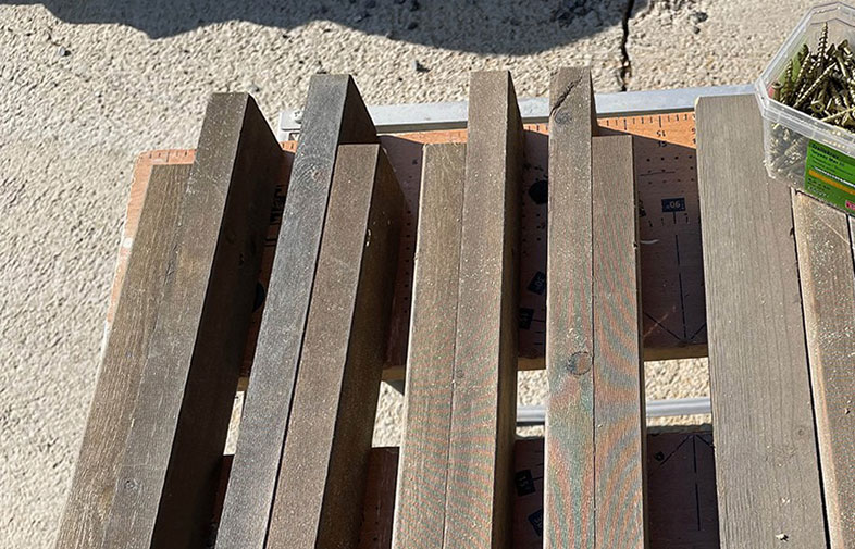 Bruna träplankor på ställningsbord med linjalmarkeringar, burk med skruvar i hörnet.