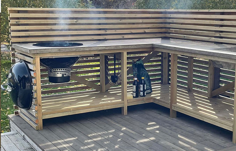 L-formad utehörna i ljust trä med svart inbyggd grill, rök och ölflaskor.