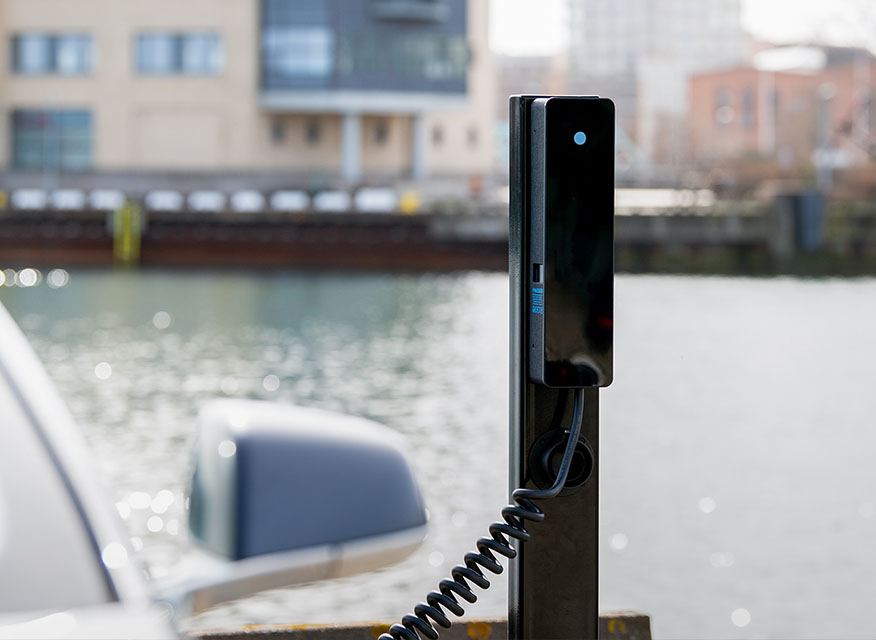 Svart elbilsladdstolpe med ihopkryllad kabel vid kajkant, silverbilsspegel och byggnader.