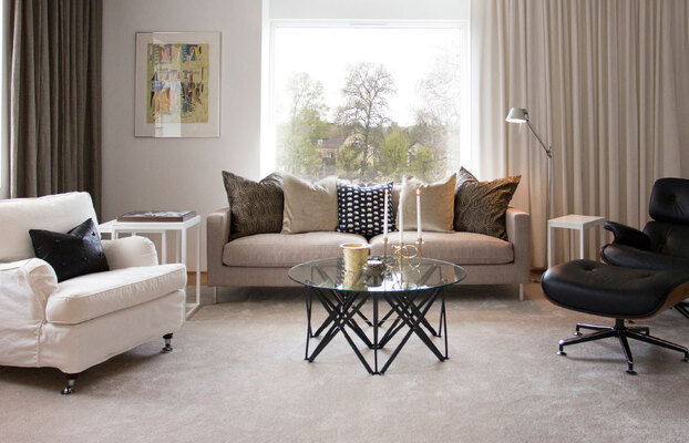 Ljust vardagsrum med beige soffa, vit fåtölj, svart vilstol, geometriskt glasbord och stort fönster