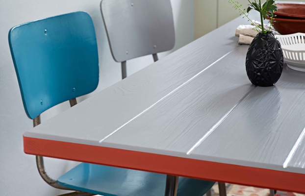 Grått bord med röd kant, två stolar (turkos och ljusgrå), svart vas med grönt blad.