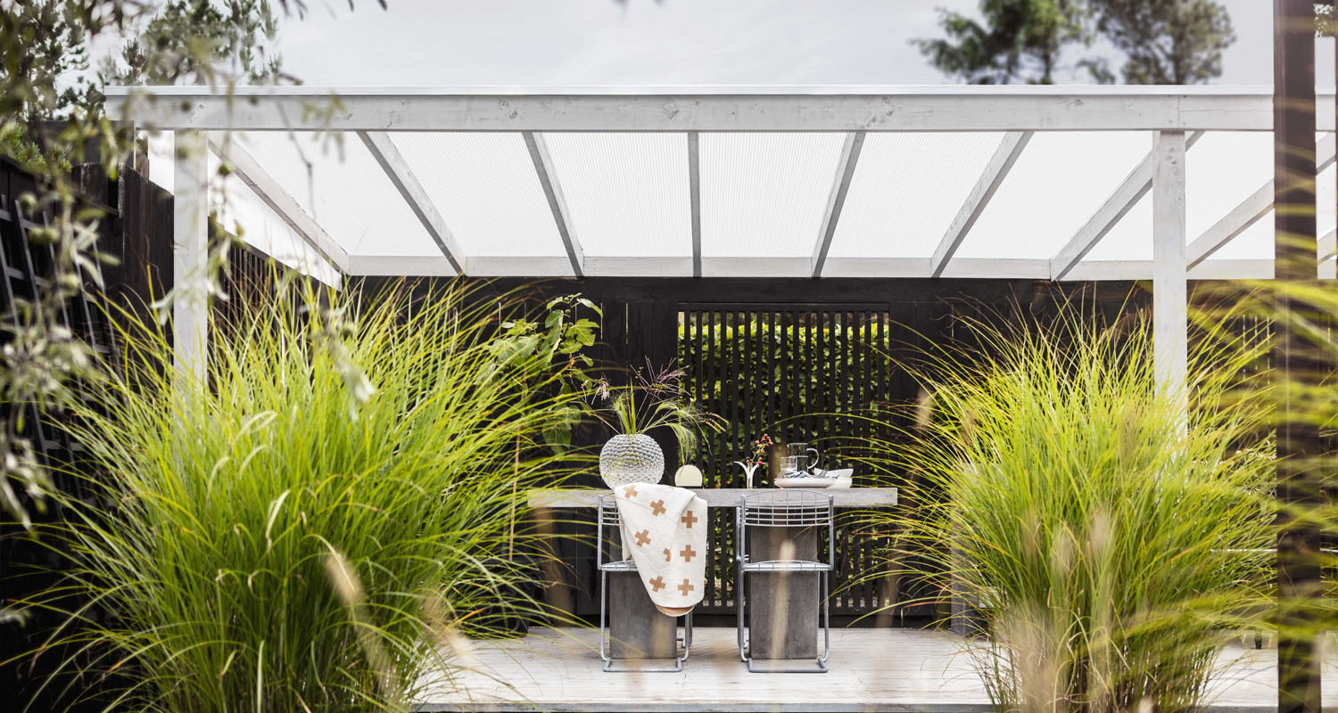 Uteplats med vitt pergolatak, betongbord och stålstolar, omgiven av frodigt grönt prydnadsgräs