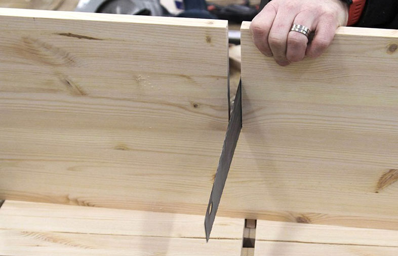 Hand med ring trycker träbit mot metallisk sågklinga på bordssåg mellan ljusa plankor