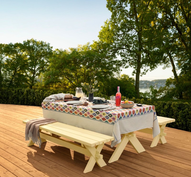 Bord dukat med mönstrad duk, rosa flaska och träbänkar på utealtan mot sjö och skog