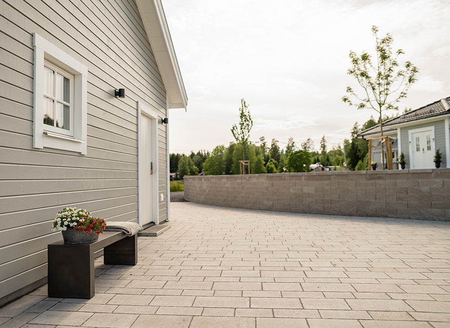 Grå trähusfasad med vit dörr, bänk med röd-vit blomkruka på ljus stenläggning.