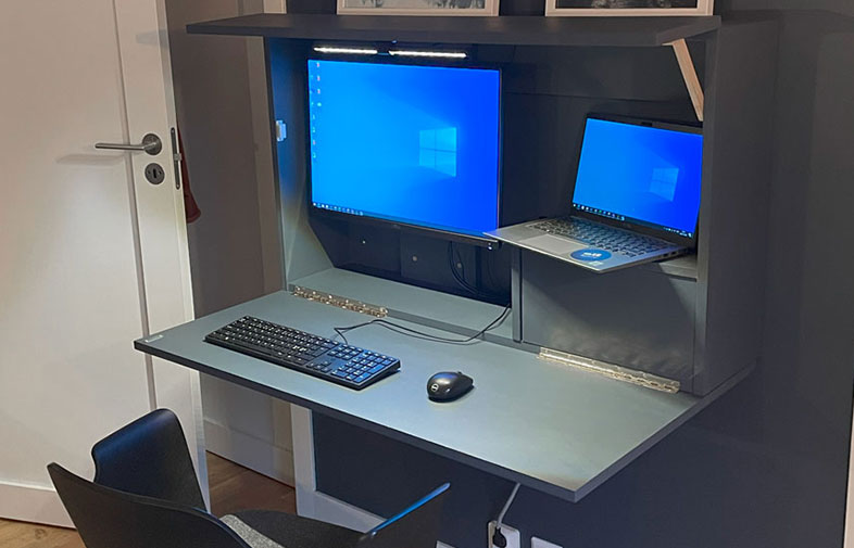 Väggmonterat mörkgrått fällbart bord med två blå skärmar, tangentbord, mus och svart stol