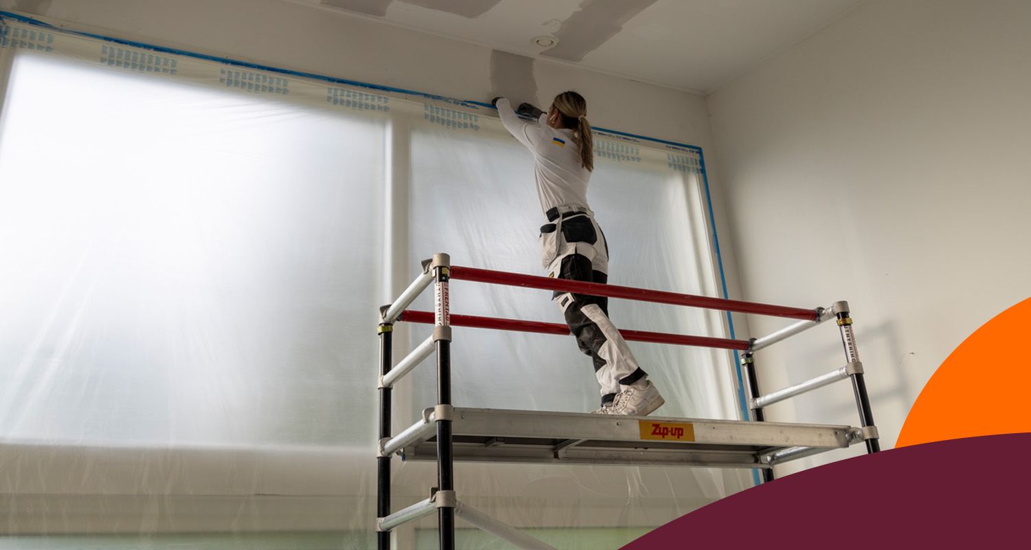 Målare i vit klädsel på ställning med röda räcken spacklar vägg vid plasttäckta fönster.