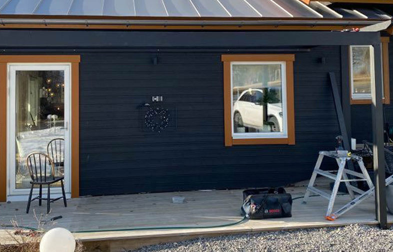 Svart träfasad med orange fönsterlister, stol och verktyg på träaltanen, vit bil reflekteras.