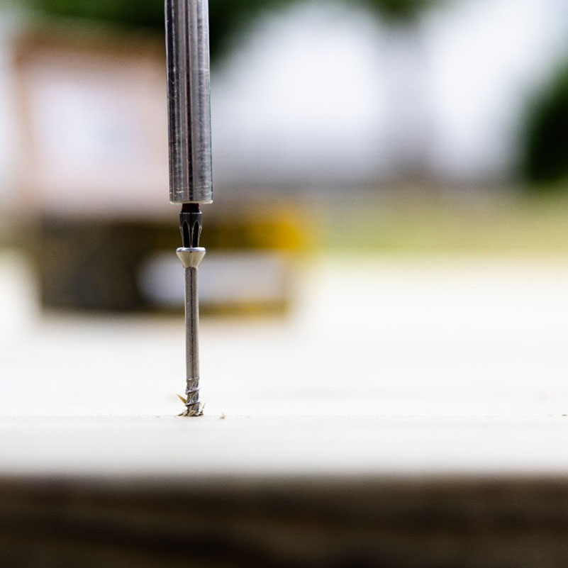 Grå metallbits driver silvrig skruv i ljust trä, suddig gulvit bakgrund.