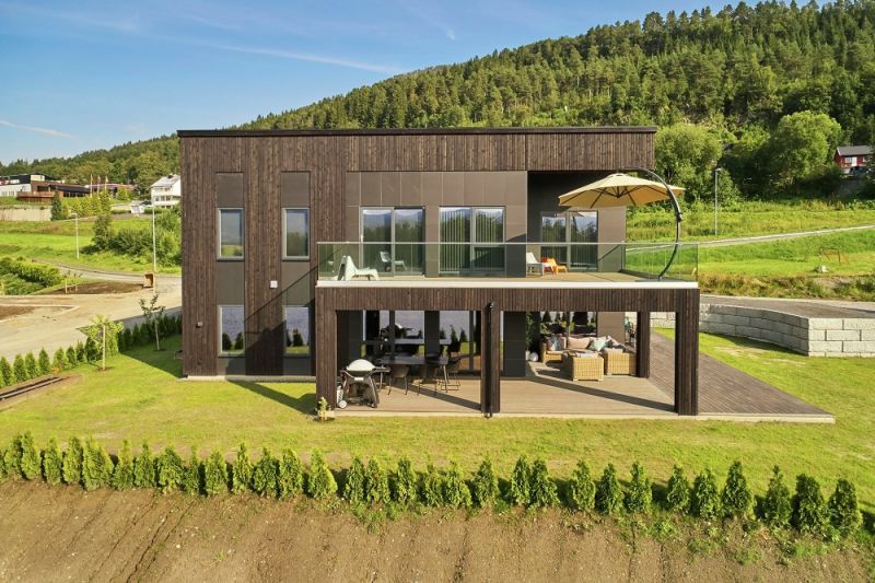 Modernt tvåvåningshus i mörkt trä med glasräcke, parasoll och skogsbacke.