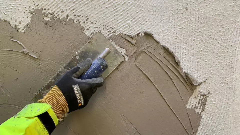 Svartgul handske med blå murslev sprider beige puts på vit, strukturerad vägg.