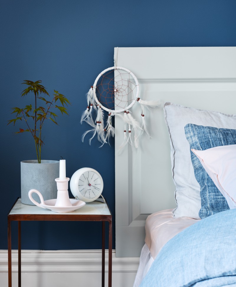 Drömfångare med vita fjädrar på vit sänggavel mot blå vägg, sängbord med växt, klocka och ljus.