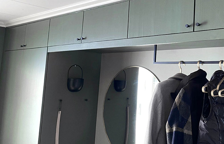 Entréhall med gröna skåp, rund väggspegel och mörka jackor på klädstång