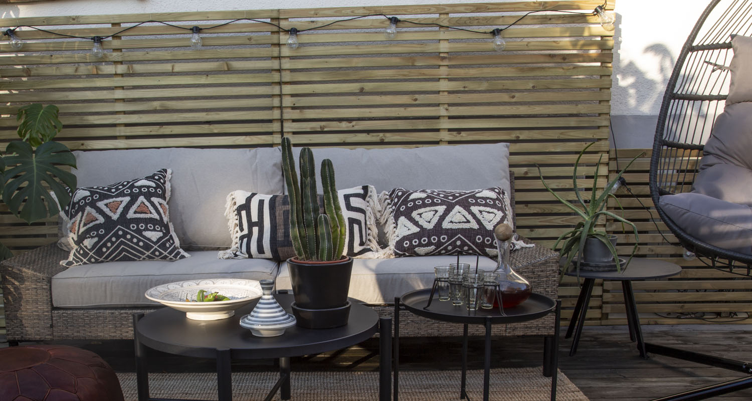 Uteplats med beige rottingsoffa, svartvita mönstrade kuddar, kaktus på svart bord framför träspaljé.