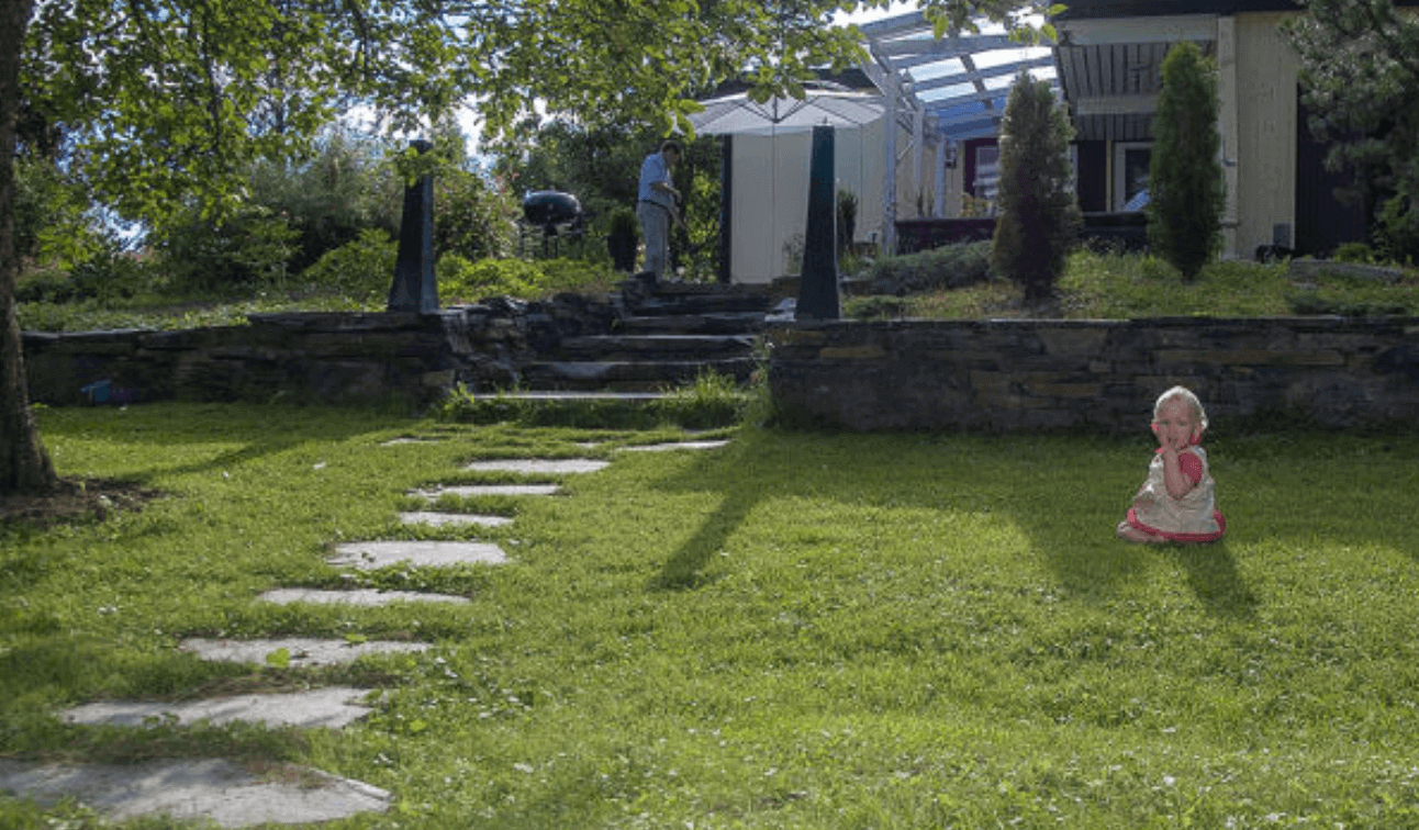 Barn i rosa klänning sitter på grön gräsmatta med stenbelagd gång mot hus med vit parasoll