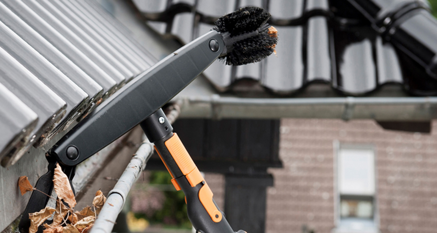 Svartorange teleskopborste rensar torra löv ur en grå plåtränna under mörkgrå takpannor.