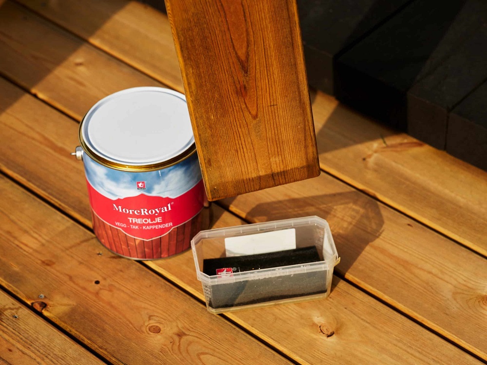 Röd-blå MoreRoyal träolja, plastask med svamp och mörkbehandlad träplanka på ljus trall