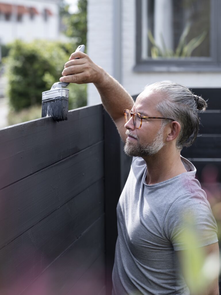 Person målar svart staket med pensel i en solig trädgård.