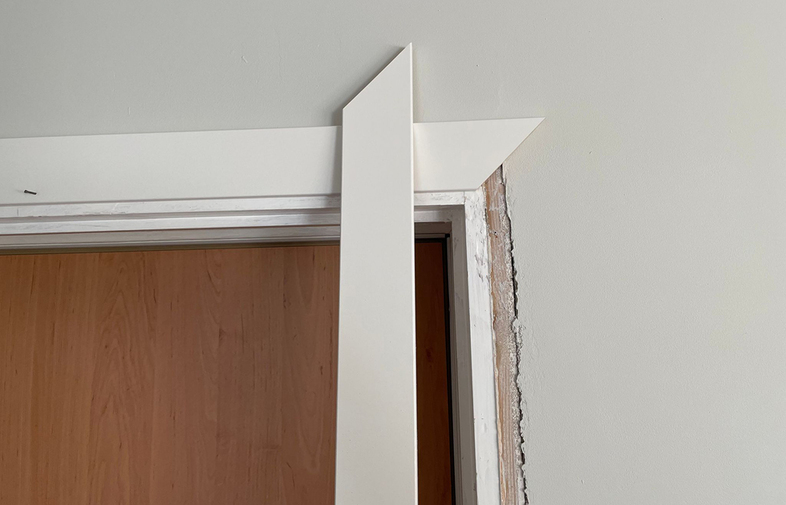 Närbild på vit dörrkarm med snedställd list mot ljusgrå vägg och brun dörr.