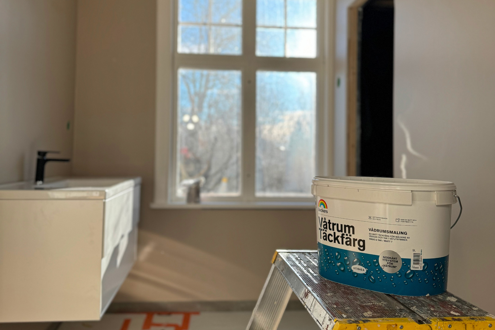 Målarfärgshink på stege i ljust badrum med beige väggar och fönster