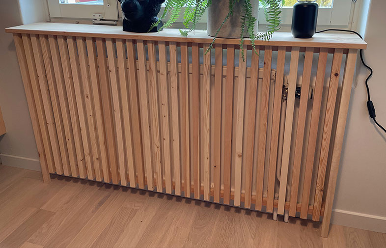 Ljus träskärm med vertikala spjälor framför vitt element, grön ormbunke i kruka ovanpå