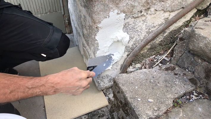 Person som spacklar en sprucken stenmur med vitt spackel vid grå trappsteg utomhus.