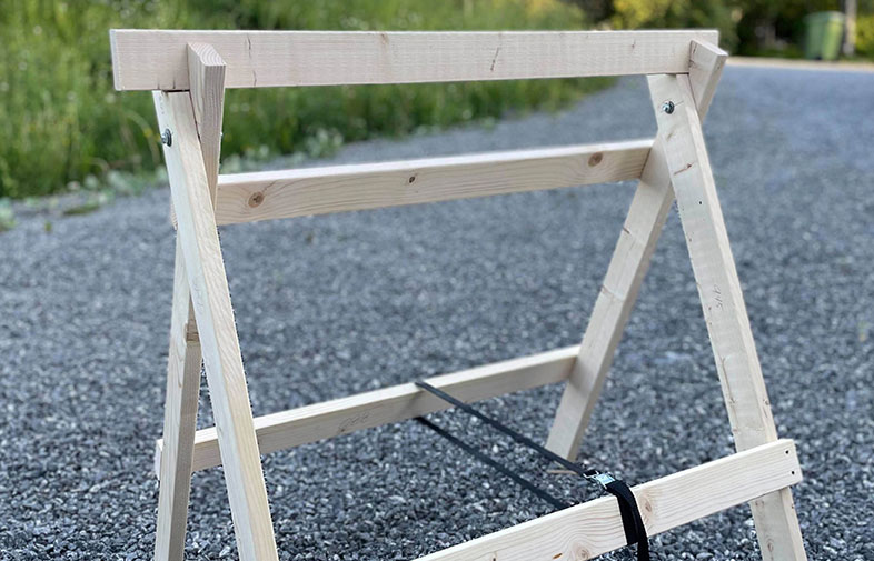 Ljust obehandlat träbocksställning med svart spännband på grusväg utomhus, grön bakgrund.