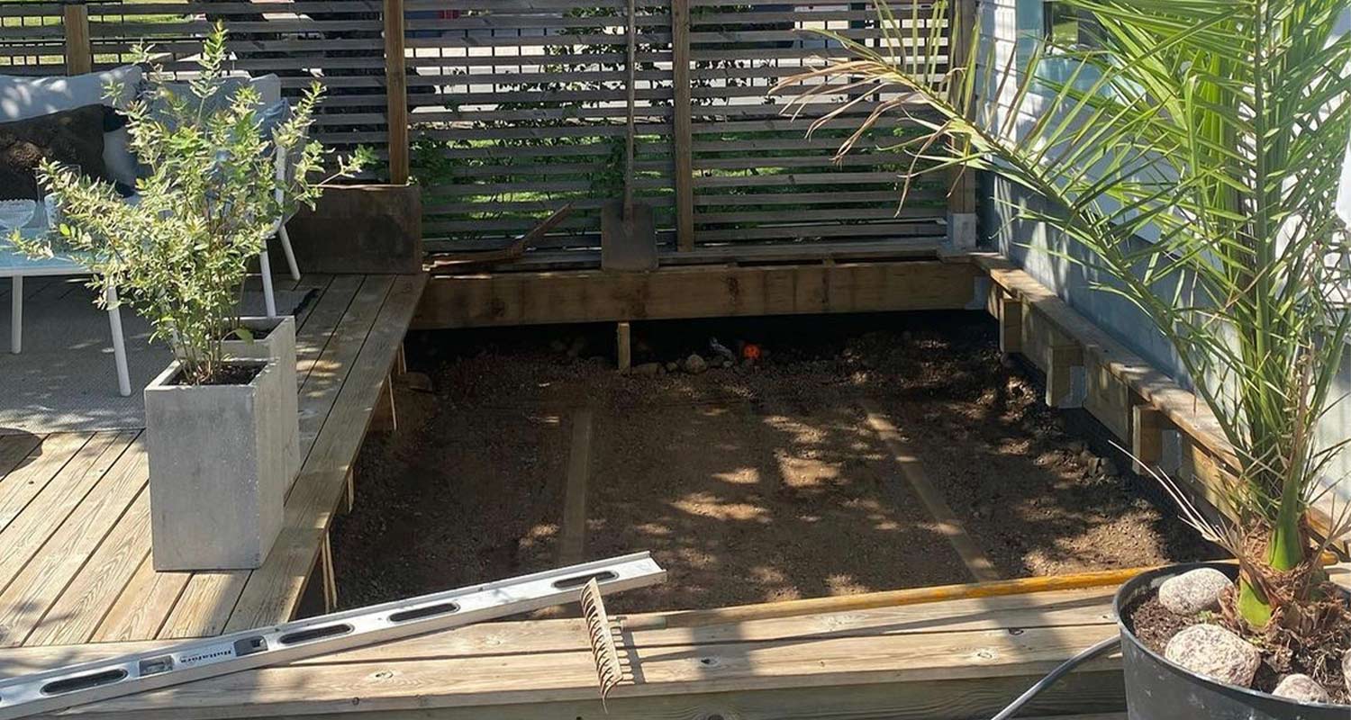 Delvis uppgrävt trädäck med vattenpass, kratta, betongkruka med grön växt och palm.