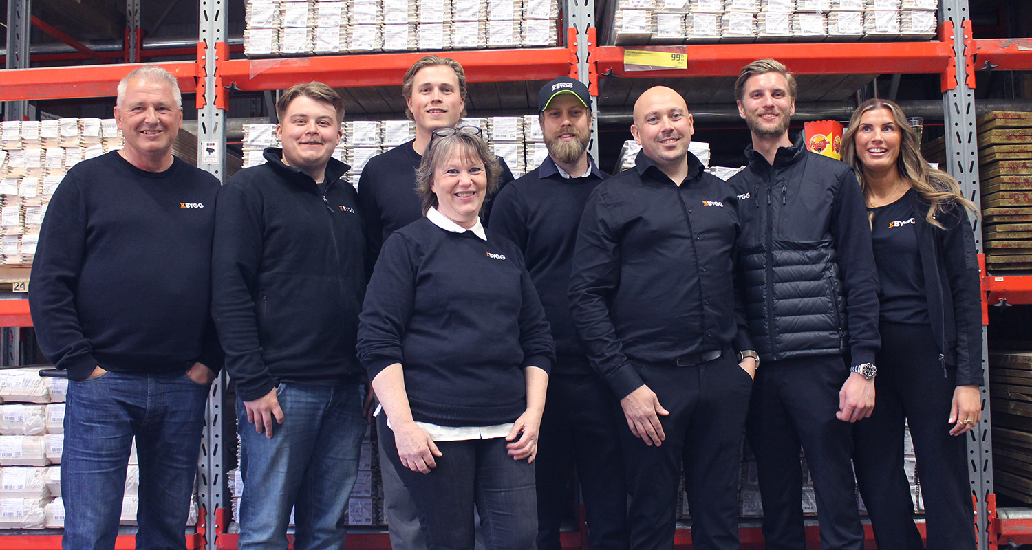 Grupp på åtta i svarta byggkläder framför röda metallhyllor med staplade kartonger