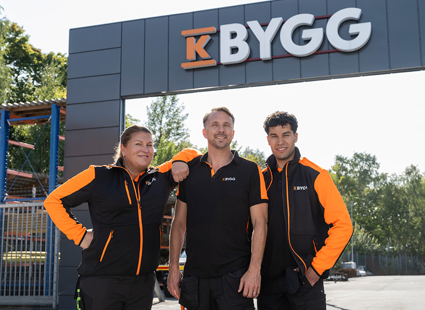 Tre byggarbetare i svart och orange står under en “BYGG”-skylt vid montageställningar.