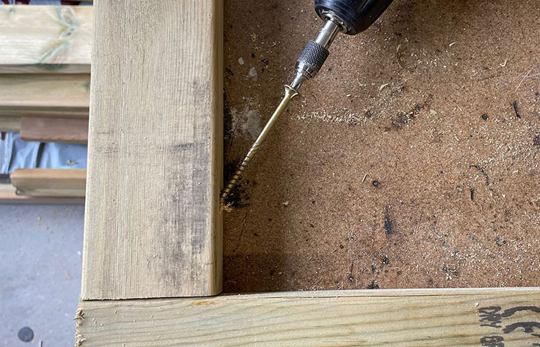 Närbild på tryckimpregnerade träreglar i hörn, borrmaskin med gyllene träskruv och sågspån.
