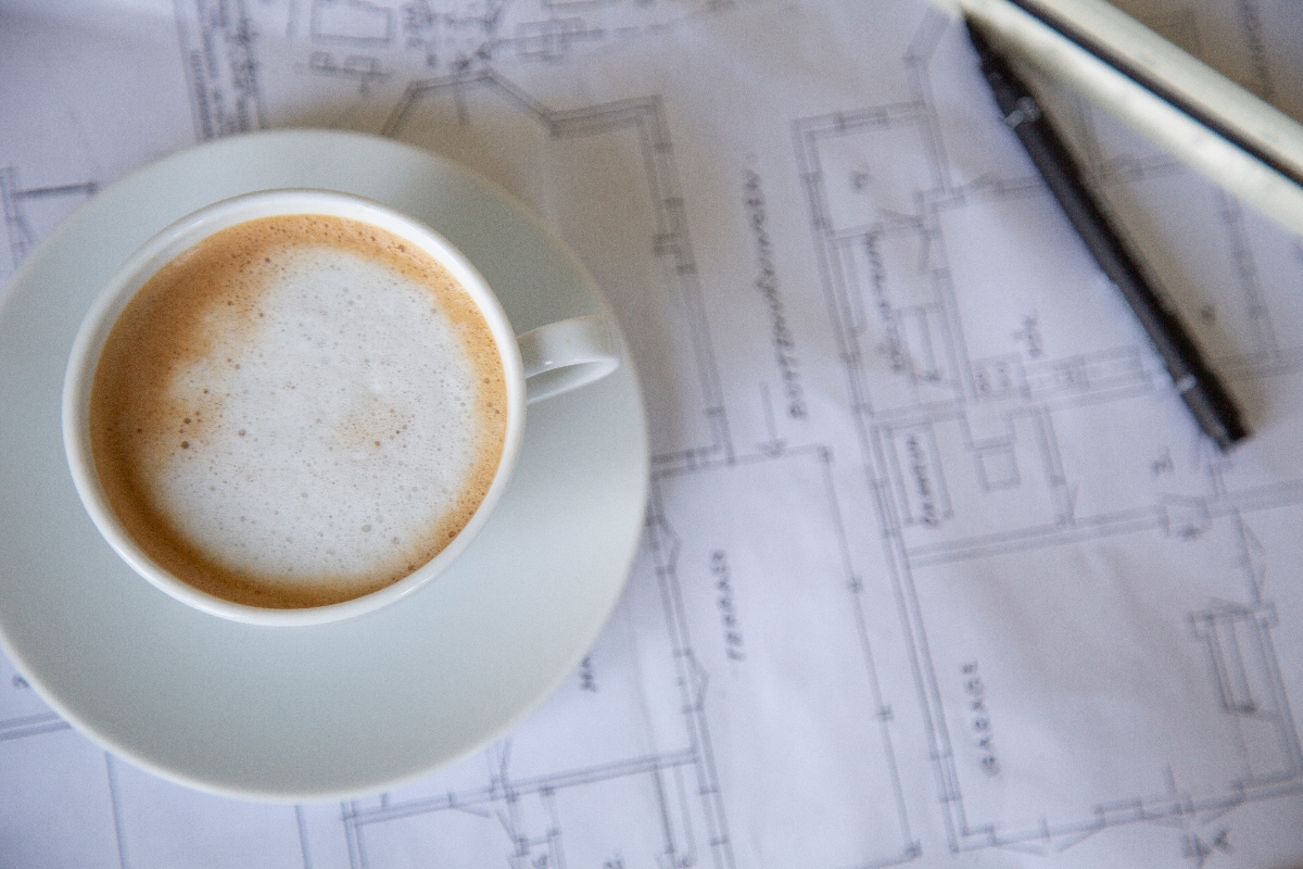 Vit kaffekopp med mjölkskum på arkitektritning och svart penna, topvy.