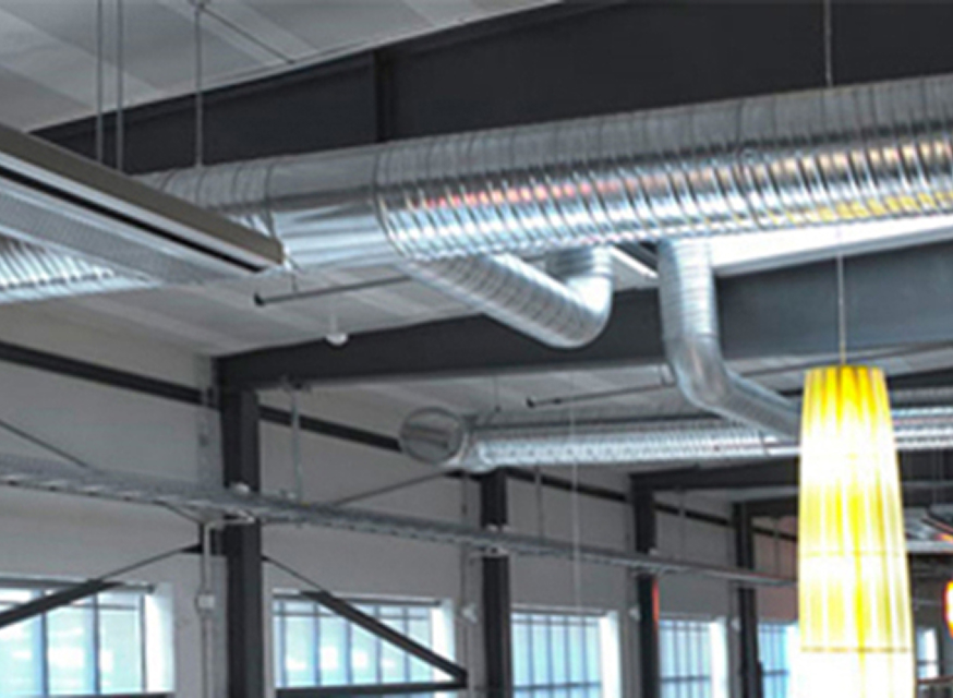 Exponerat industriloft med silvriga ventilationsrör, svarta balkar och gul pendellampa