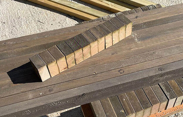 Mörkbruna träbrädor staplade på sand, med ljusa träblock i rad på översta brädan.