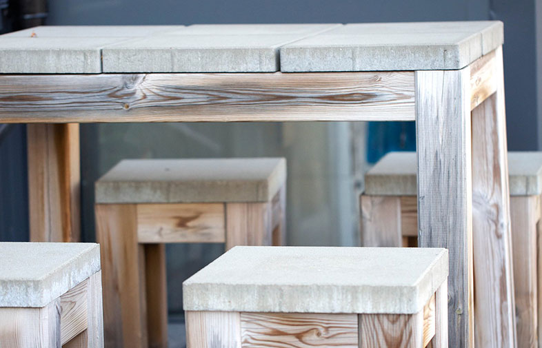 Högt bord och fyra pallar i ljus trä med grå betongliknande skivor, utomhusmiljö.